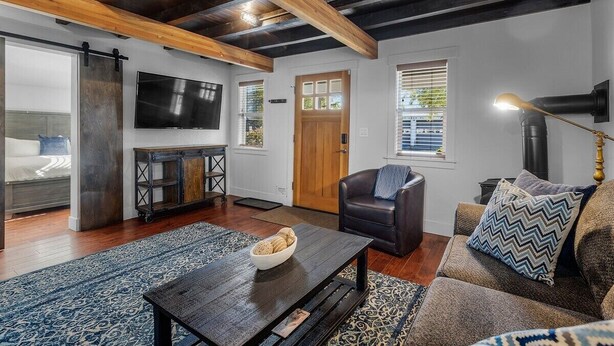 A modern living room with a wooden ceiling, a black leather sofa, a patterned rug, and a television mounted on the wall. - Bluebird Day - Lakewood Cottage in Bend with 2 beds, perfect for 4 guests. (Bend)