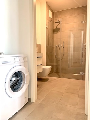 Une salle de bain moderne équipée d’un lave-linge, de toilettes et d’une douche à parois de verre.