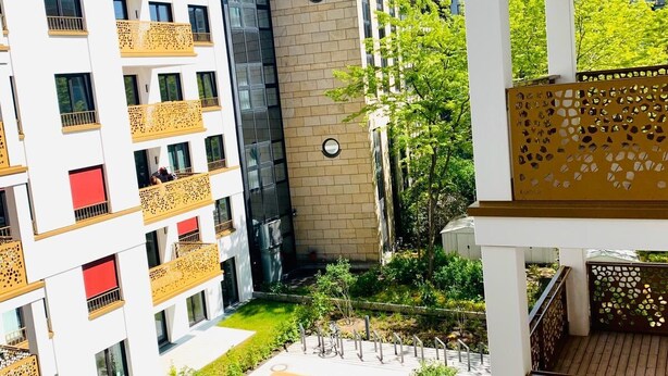 Un immeuble d’appartements moderne doté de nombreux balcons, d’une cour intérieure verdoyante et d’un mélange de bâtiments résidentiels et commerciaux en arrière-plan.