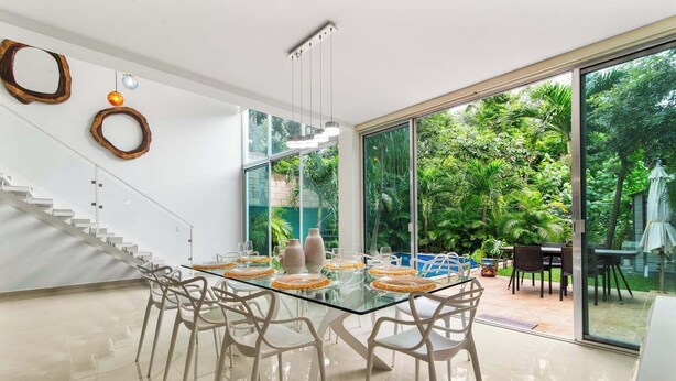 Un espace repas moderne avec une table en verre et des chaises blanches, entouré de grandes fenêtres offrant une vue sur une végétation luxuriante.