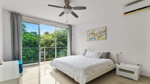 Une chambre à coucher avec un lit, un ventilateur de plafond, une fenêtre avec des rideaux, une table de chevet et un tableau accroché au mur.