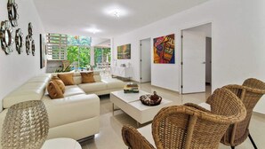 Un salon moderne avec un canapé blanc, des fauteuils en osier et une table basse. Les murs sont ornés de tableaux et, par la fenêtre, on aperçoit un paysage verdoyant.