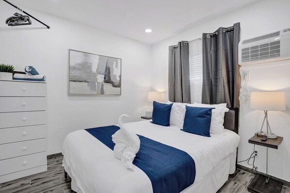 A hotel room with a bed, a dresser, a painting, a window with curtains, and an air conditioning unit.