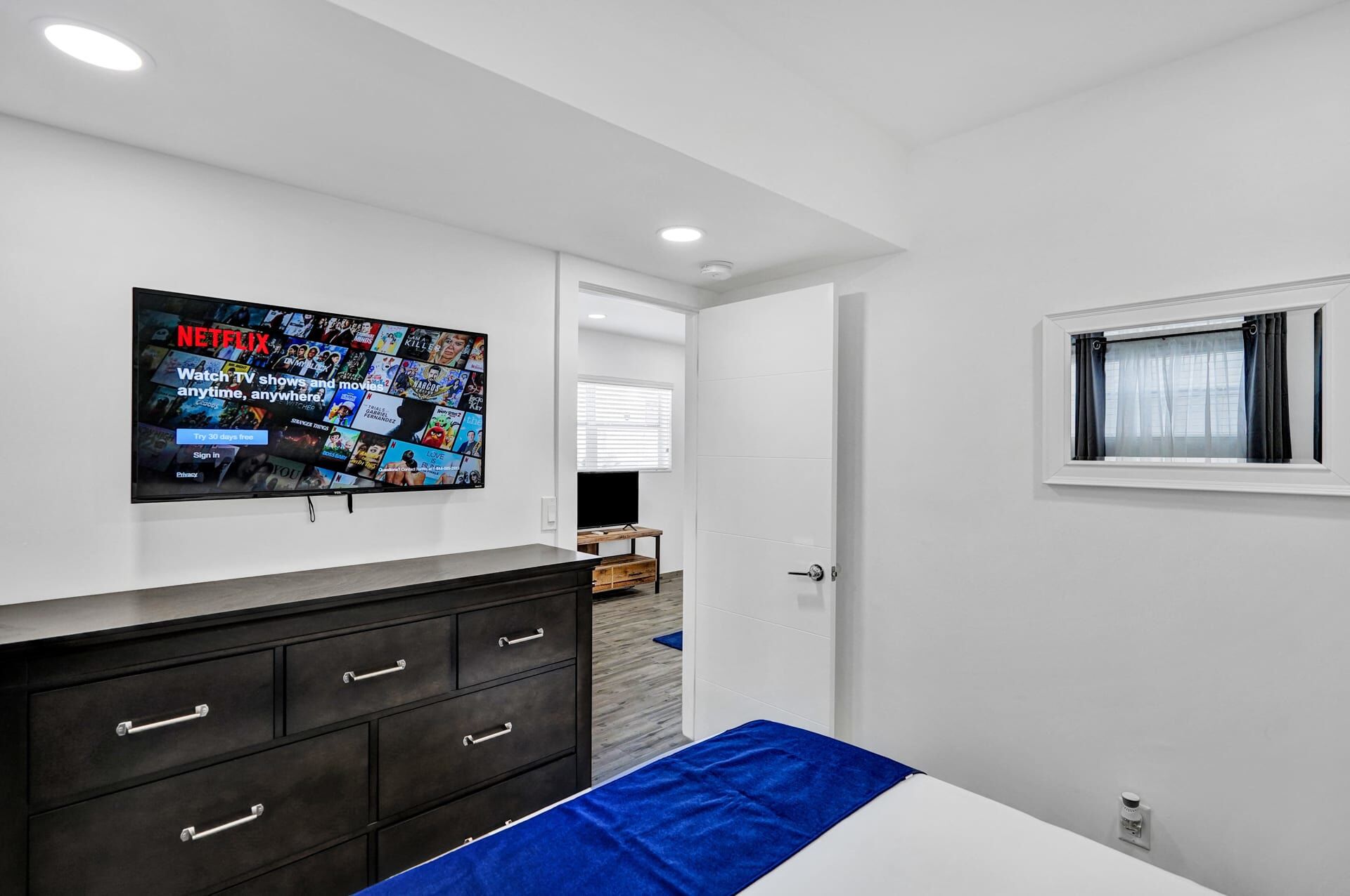 Un dormitorio moderno con un televisor de pantalla plana grande montado en la pared, mostrando la interfaz de Netflix. Hay un aparador de madera con cajones y una colcha azul.