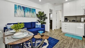 Una sala de estar moderna con un sofá azul, una mesa de comedor para dos personas y una cocina con armarios blancos y una cafetera.