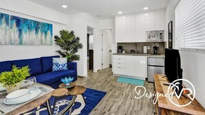 Una sala de estar moderna con un sofá azul, una mesa de centro y un comedor con una mesa y sillas de madera. Se observa una cocina con armarios blancos y un microondas.