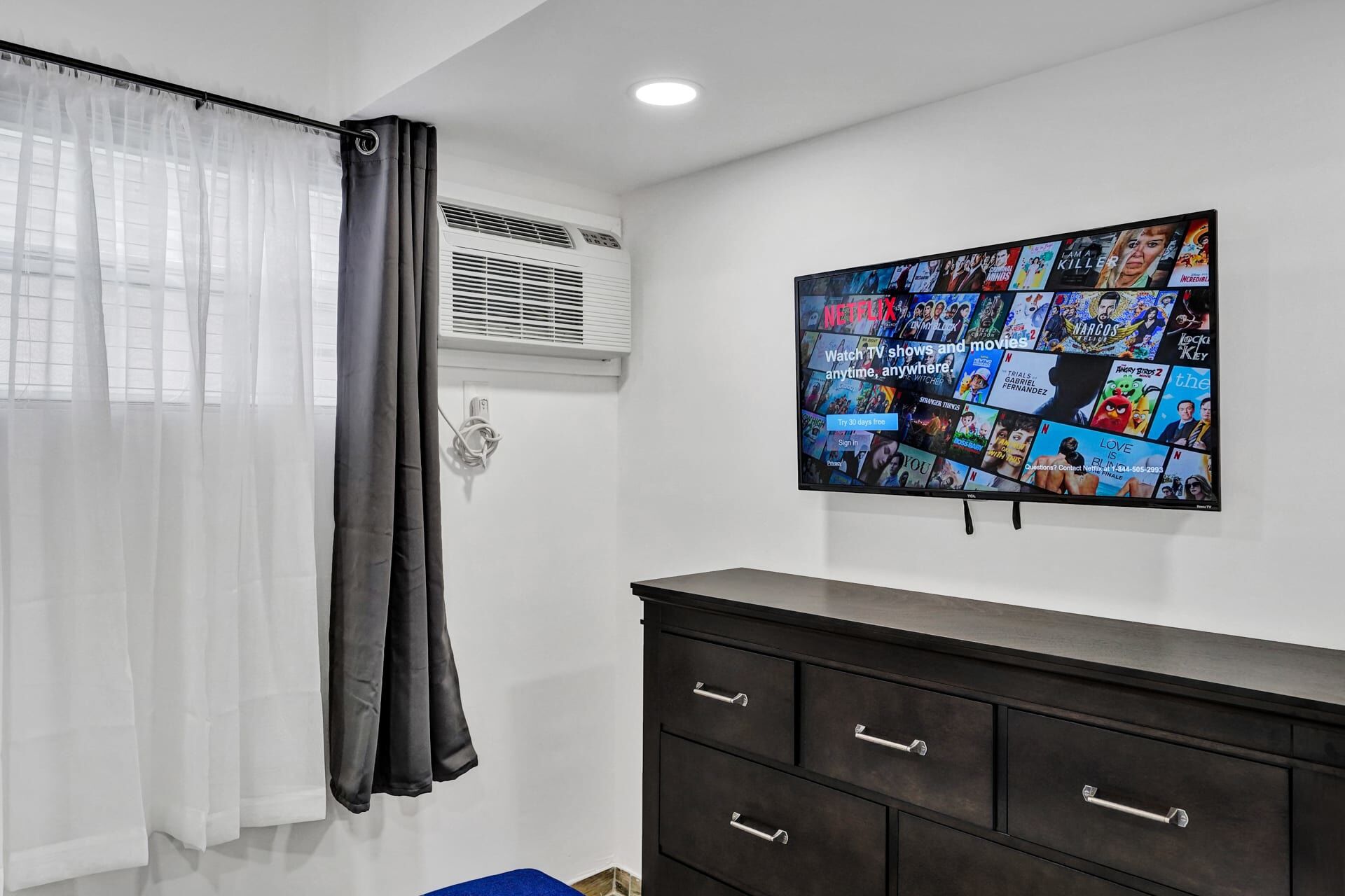A room with a large TV displaying various movie and TV show posters, a dark-colored dresser with drawers, and a window with sheer curtains.