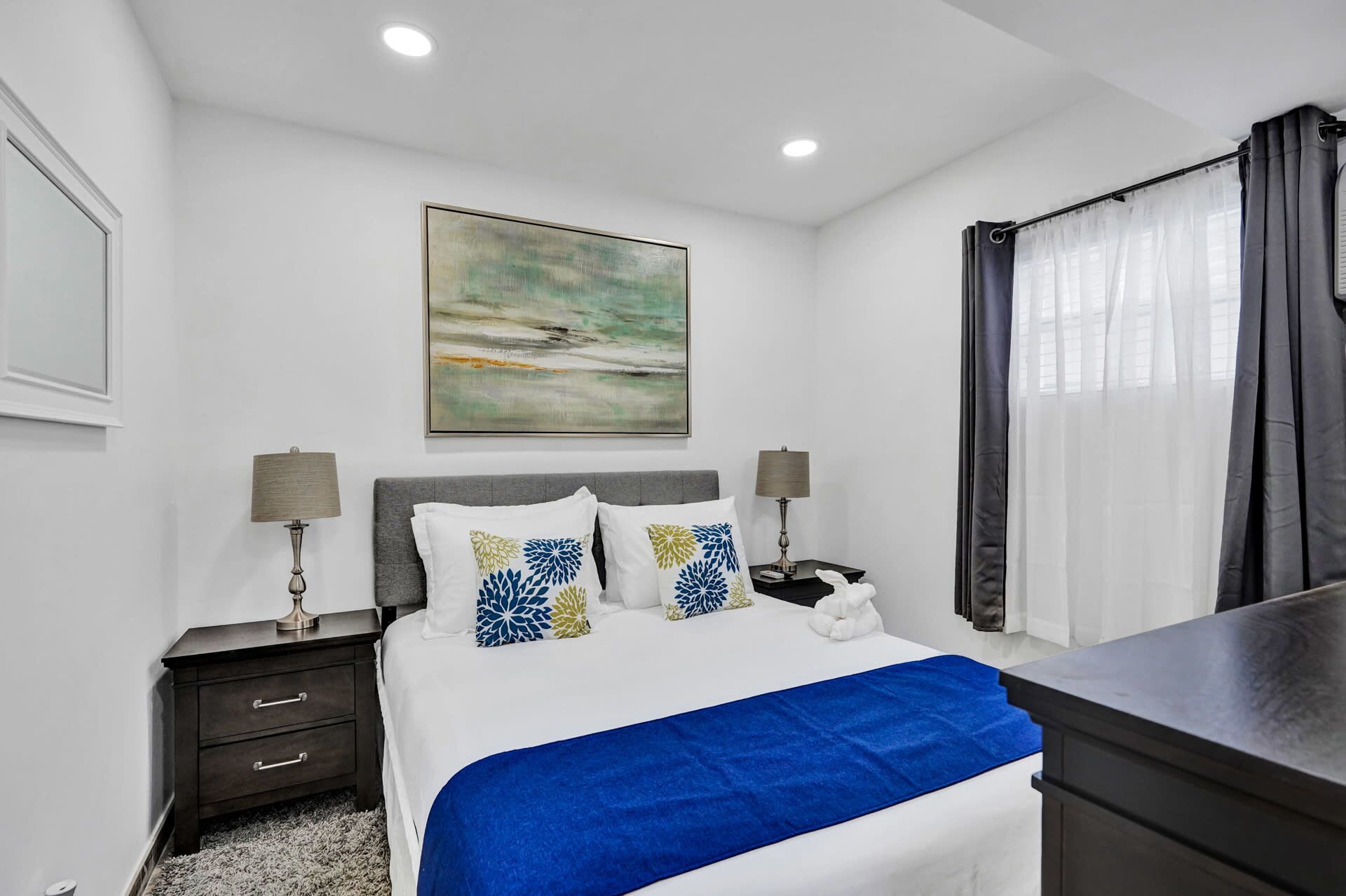 A hotel room with a bed, two bedside lamps, a nightstand, a desk, a chair, a window with curtains, and a painting on the wall.