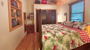 Ein Schlafzimmer mit Bett, Holzboden, Deckenventilator, Fenster mit Jalousien und Regal mit gerahmten Bildern.