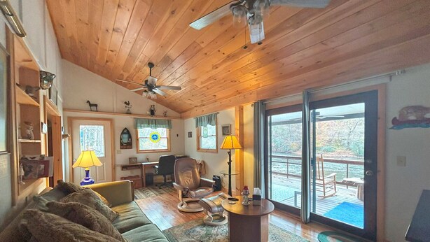 Ein gemütliches Blockhaus-Interieur mit Holzbalkendecke, Deckenventilator, einer Couch, einem Couchtisch, einem Schreibtisch und Blick auf einen See durch große Schiebeglasfenster.