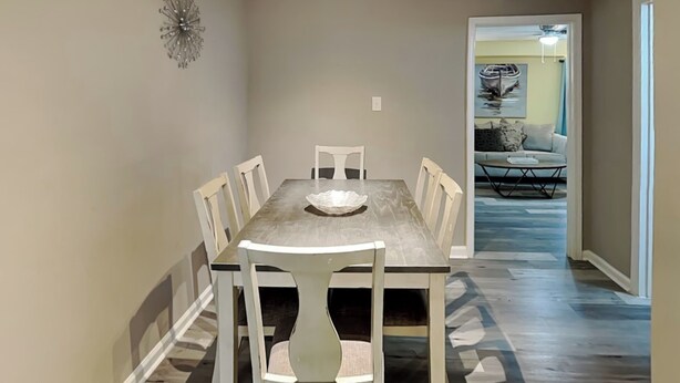 Un comedor con una mesa rectangular y seis sillas, una lámpara de araña, y un pasillo que conduce a una sala de estar con un sofá y una mesa de centro.