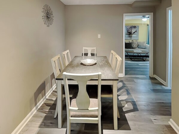 A dining area with a rectangular table and six chairs, a chandelier, and a hallway leading to a living area with a couch and a coffee table. - Pleasure Pier Retreat #3 | Fenced Backyard & Great Location (Galveston)