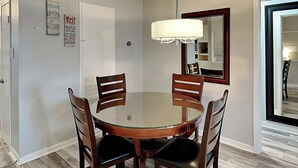 Een eetruimte met een ronde glazen tafel, vier houten stoelen, een kroonluchter en een spiegel aan de muur.