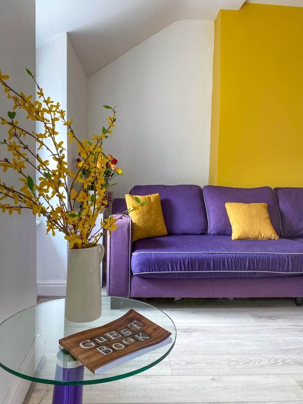 A living room with a purple sofa, yellow cushions, a glass coffee table, and a vase with yellow flowers.