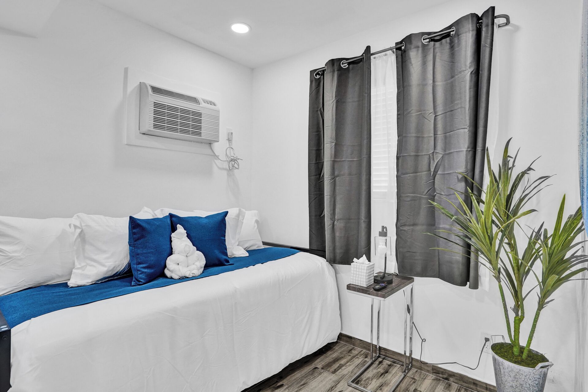 A bedroom with a bed, a bedside table, a potted plant, and a window with curtains.