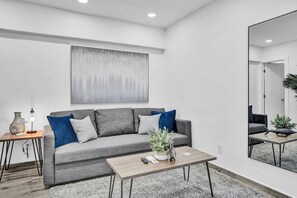 Un salon moderne avec un canapé gris, une table basse, une table d’appoint avec une lampe, et un grand tableau abstrait accroché au mur.