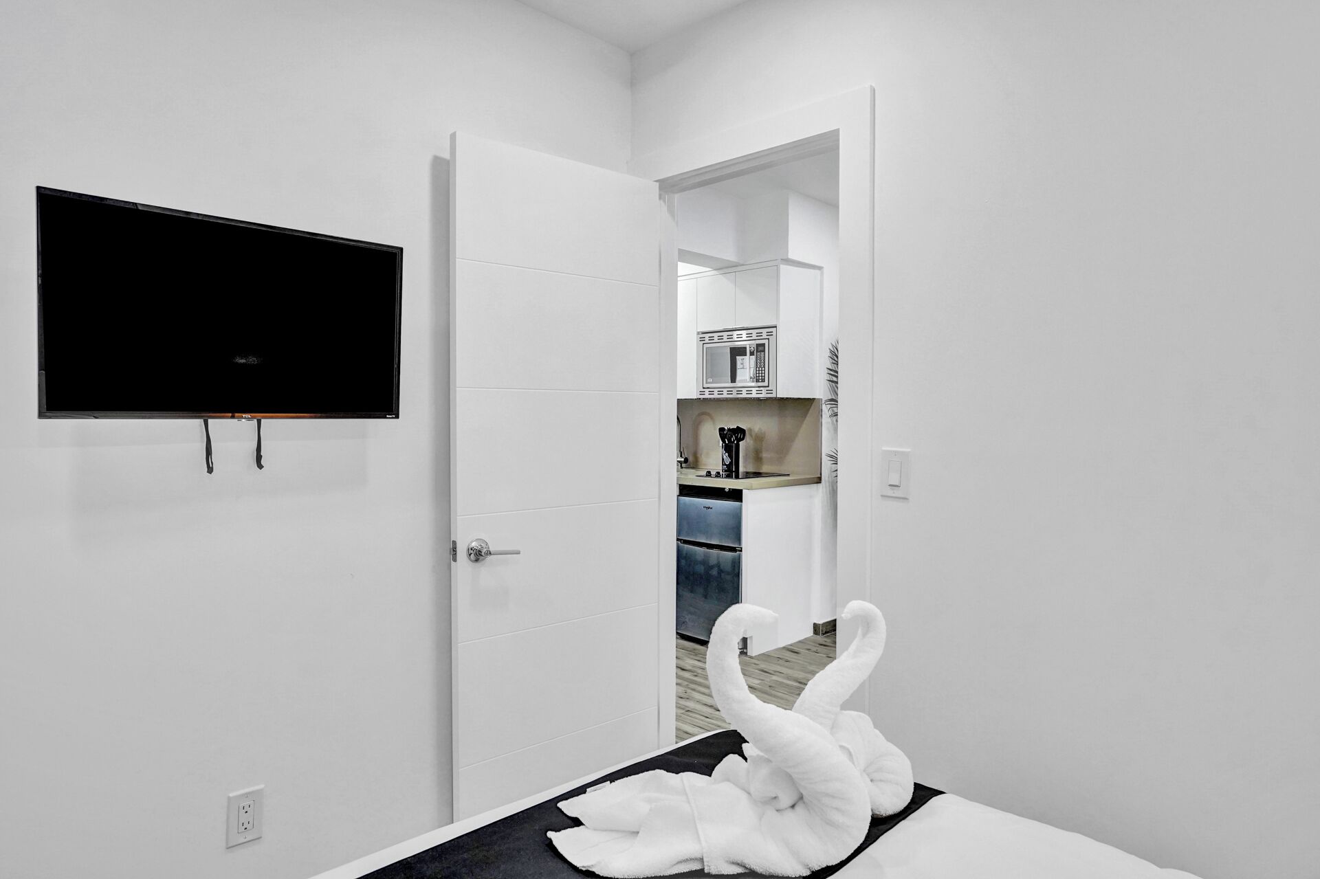 Habitación de hotel con una televisión de pantalla plana montada en la pared, una puerta blanca que da a una zona de cocina con microondas y cafetera, y una toalla con forma de cisne sobre la cama.