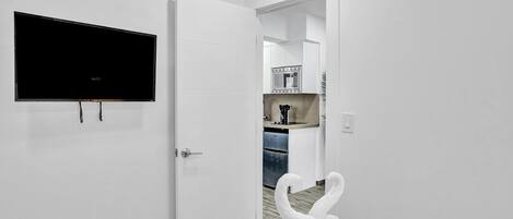 Habitación de hotel con una televisión de pantalla plana montada en la pared, una puerta blanca que da a una zona de cocina con microondas y cafetera, y una toalla con forma de cisne sobre la cama.