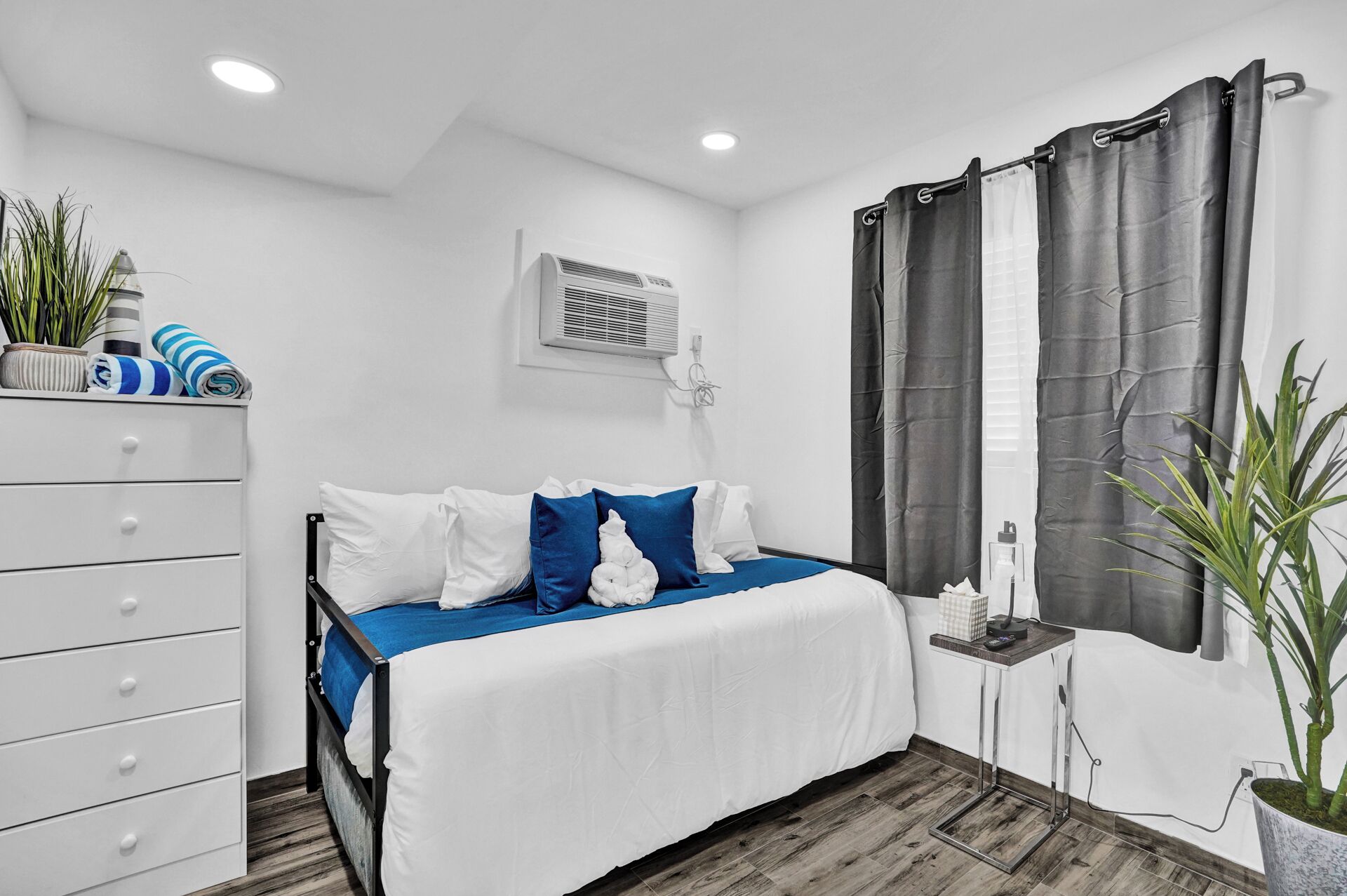 Quarto com cama, cômoda branca, aparelho de ar condicionado, janela com cortinas e um vaso com planta.