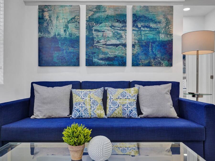 Um sofá azul com almofadas estampadas, uma mesa de centro de vidro com uma planta e objeto decorativo, e quadros abstratos na parede.