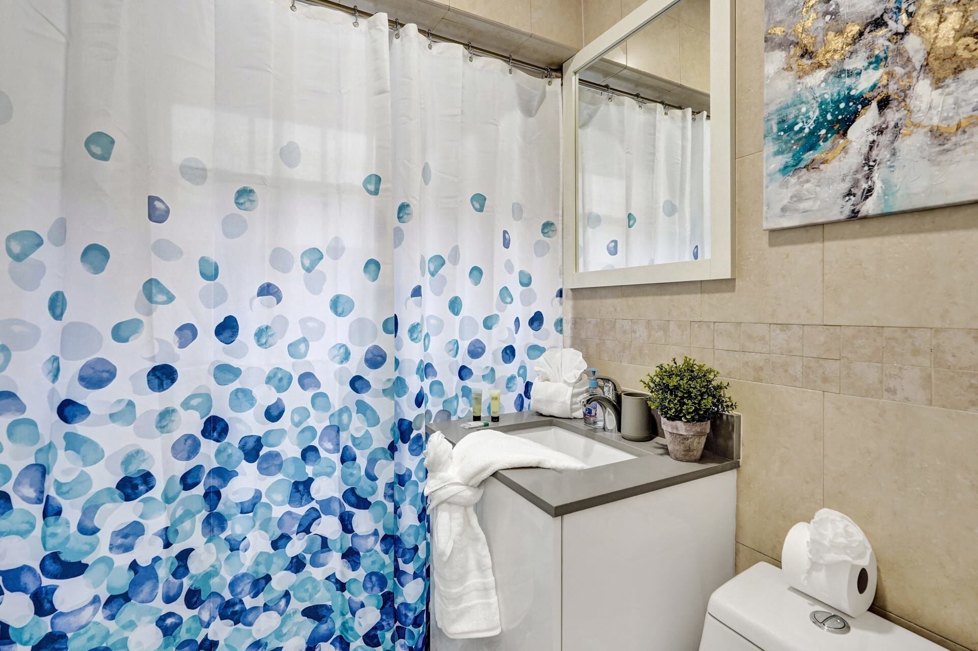 A bathroom with a shower curtain featuring blue and white abstract patterns, a sink with toiletries, a potted plant, and a toilet.