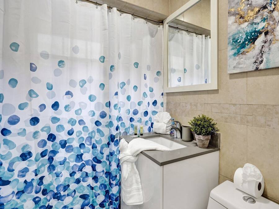 A bathroom with a shower curtain featuring blue and white abstract patterns, a sink with toiletries, a potted plant, and a toilet.