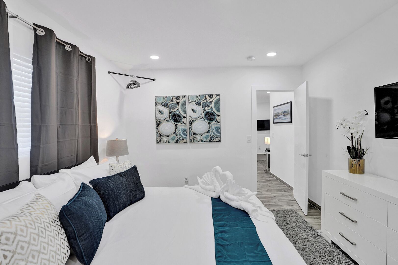 A modern bedroom with a large bed, a bedside table with a lamp, a wall-mounted artwork, and a view into the bathroom.
