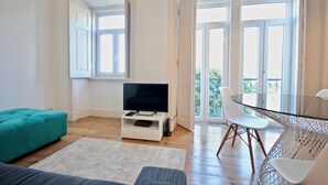 A modern living room with a television on a white stand, a teal ottoman, a glass table with white chairs, and large windows with a balcony view. - Bluebell Duplex Apartment, Anjos, Lisbon (Lisboa)