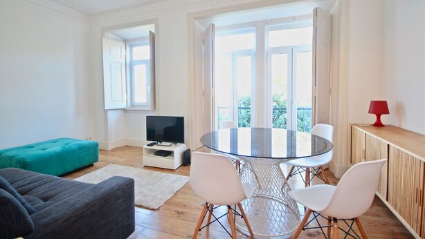 A modern living room with a glass dining table, white chairs, a teal ottoman, and a television on a white stand. - Bluebell Duplex Apartment, Anjos, Lisbon (Lisboa)