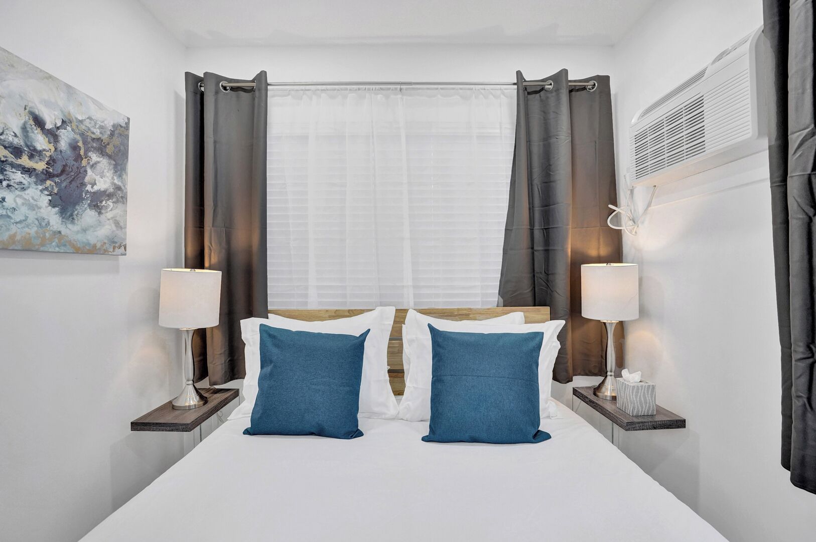 A hotel room with a large bed, two bedside lamps, a window with curtains, a painting on the wall, and an air conditioning unit.