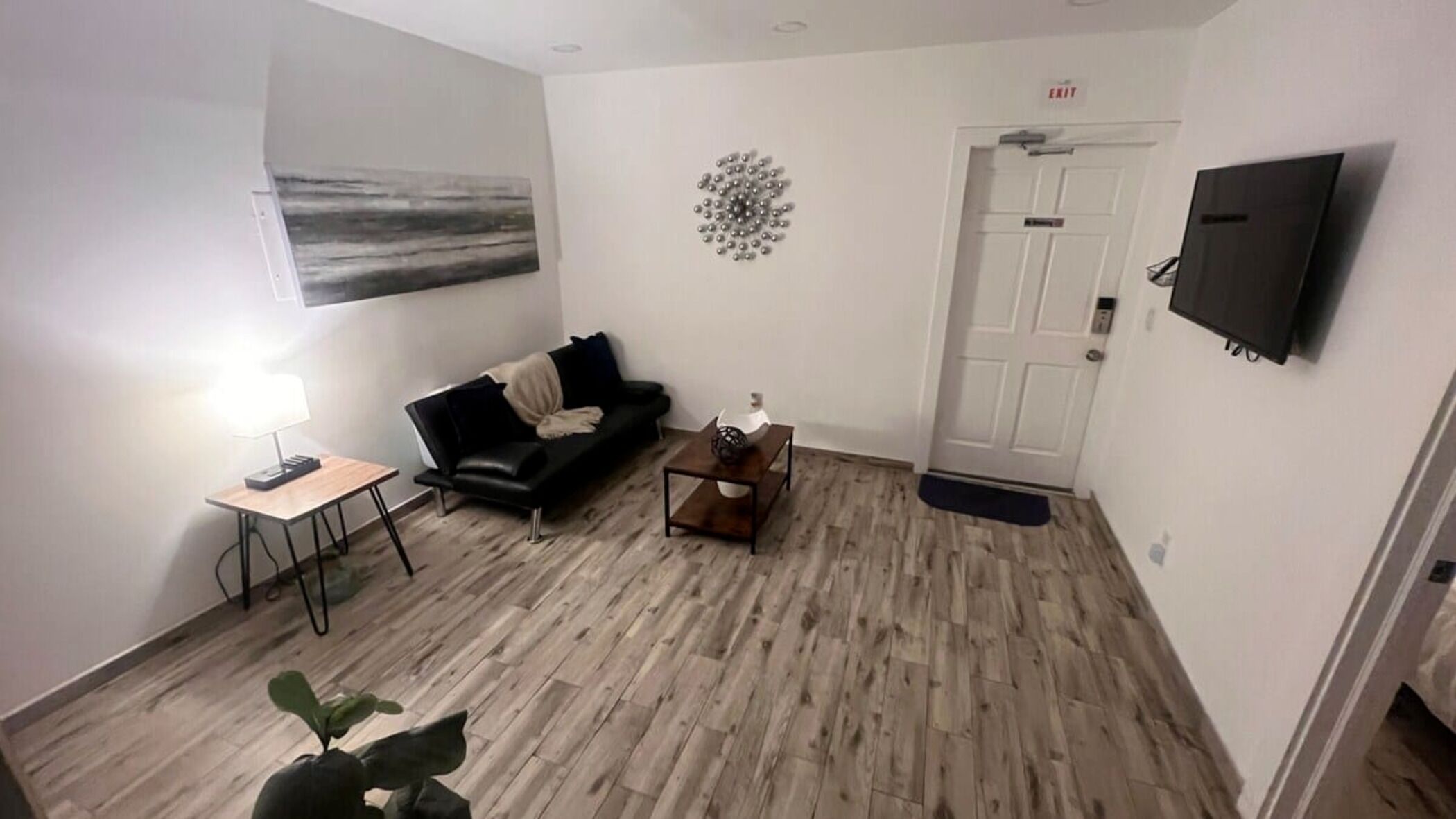 A modern living room with a black leather sofa, a wooden coffee table, a small side table with a lamp, a painting on the wall, and a flat-screen TV mounted on the wall.