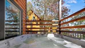 A hot tub with a wooden deck and glass walls, surrounded by trees and a cabin in the background.