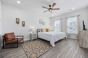 Une chambre à coucher comprenant un lit, une chaise, une table de chevet, un ventilateur de plafond et une fenêtre avec des rideaux.