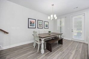 A dining area with a wooden table, white chairs, a bench, and a chandelier. There are three framed pictures on the wall.