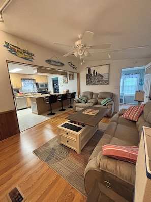 Un salon avec un canapé, une table basse et un coin repas avec des chaises. On aperçoit un ventilateur de plafond et une cuisine à l’arrière-plan.