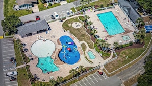 Eine Vogelperspektive zeigt ein Resort mit mehreren Swimmingpools, einem Spielbereich für Kinder und einem Parkplatz.