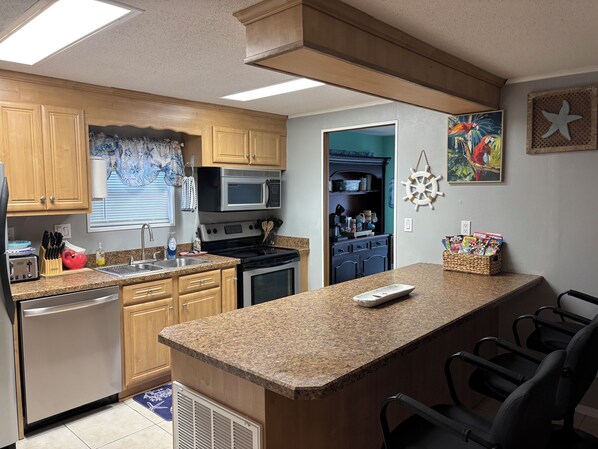 A kitchen with wooden cabinets, a microwave, stove, and a sink. There is a bar area with chairs and a remote control. - Book for your Fall Break Now!3 BR-2 B, limo-golf cart, linen, great outdoor area (Surfside Beach)