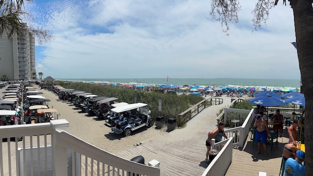 Blick auf einen Strand mit einem Parkplatz für Golfwagen, einer Holzterrasse und Strandsonnenschirmen.