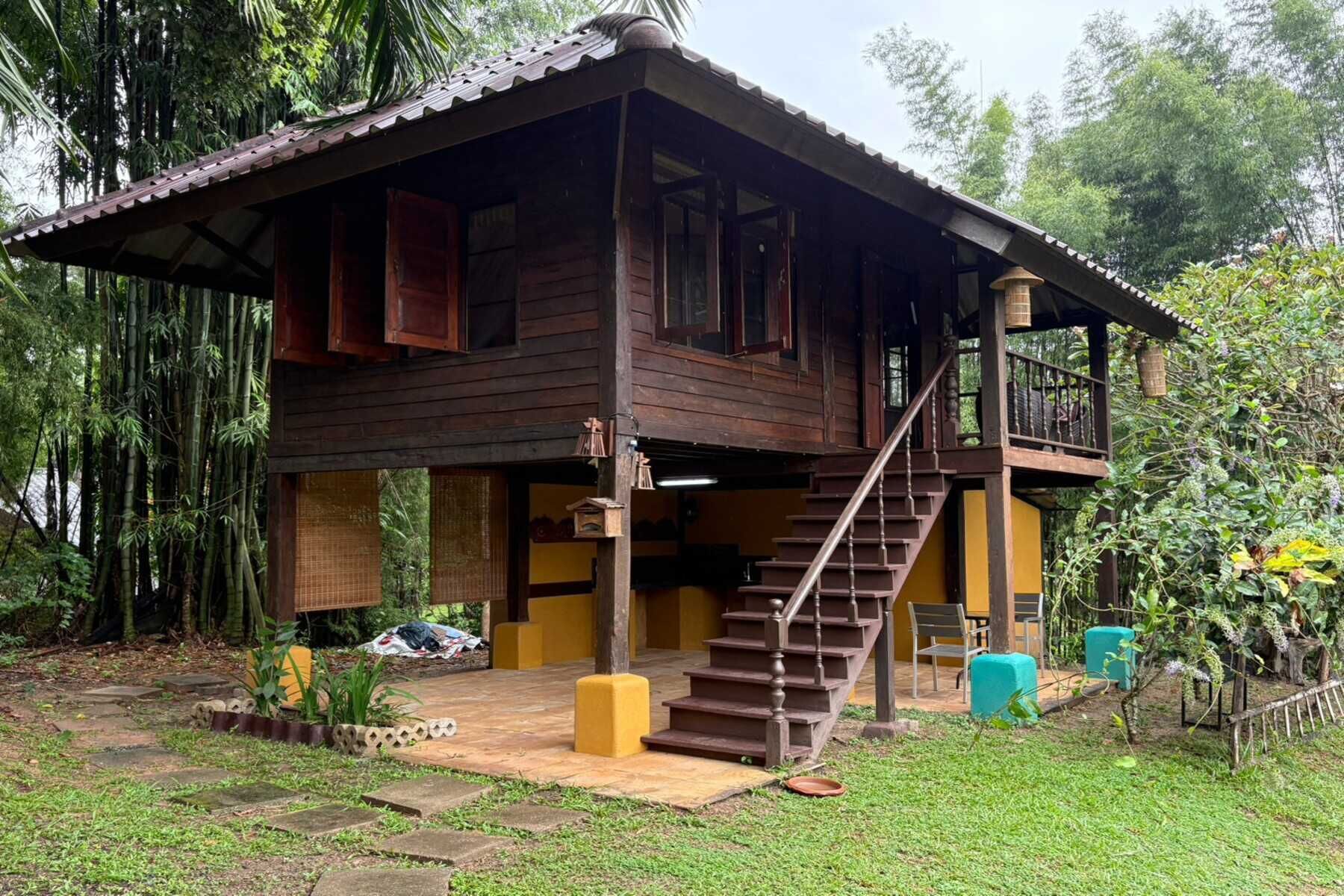 A wooden house with a staircase leading to a balcony, surrounded by greenery.