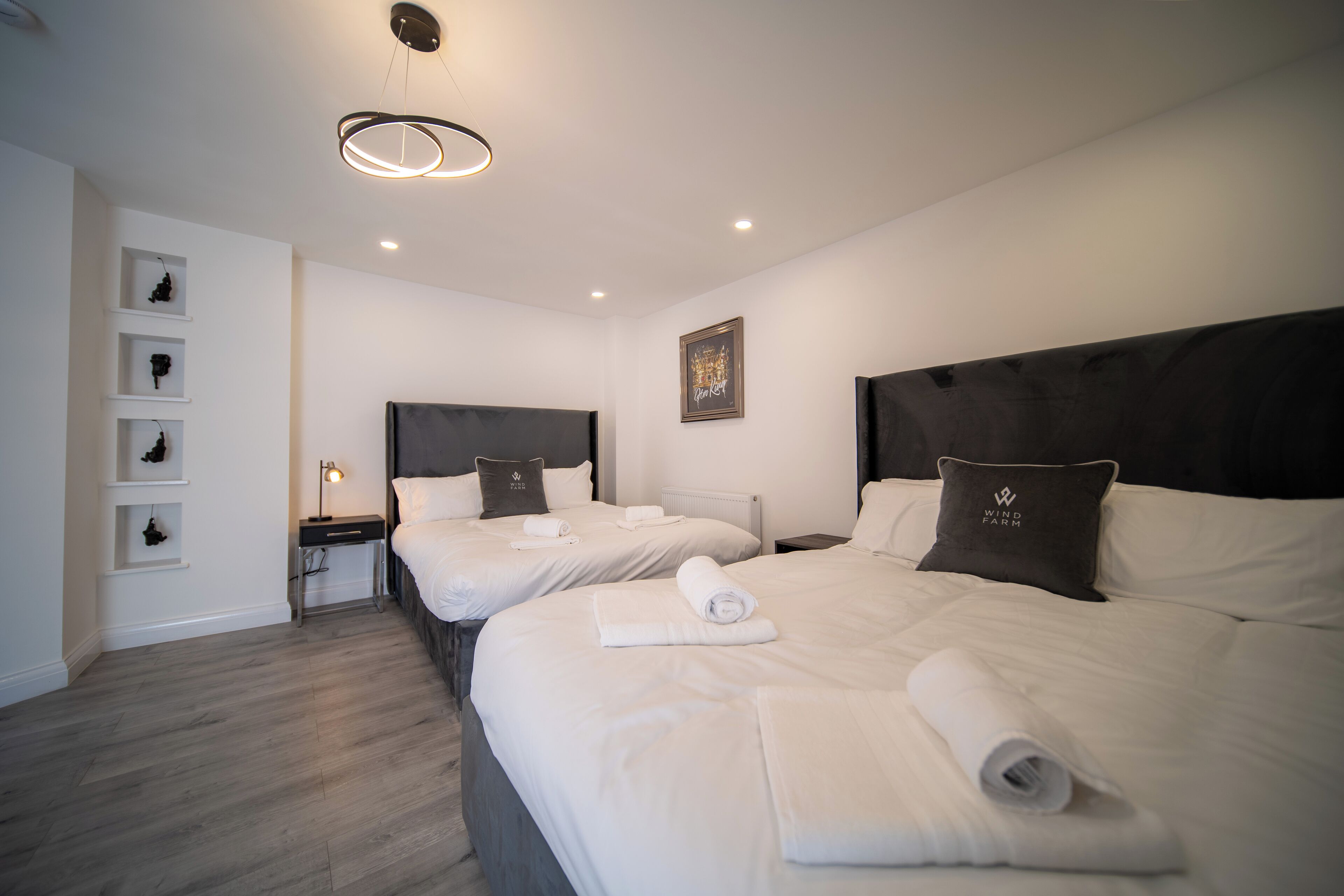 A hotel room with two beds, each with a headboard, a bedside table with a lamp, and a wall-mounted shelf with decorative items.
