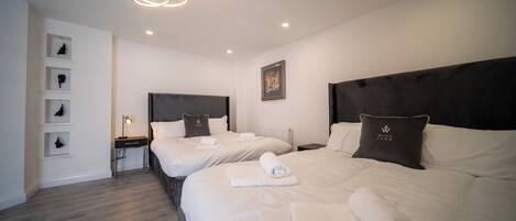 A hotel room with two beds, each with a headboard, a bedside table with a lamp, and a wall-mounted shelf with decorative items.