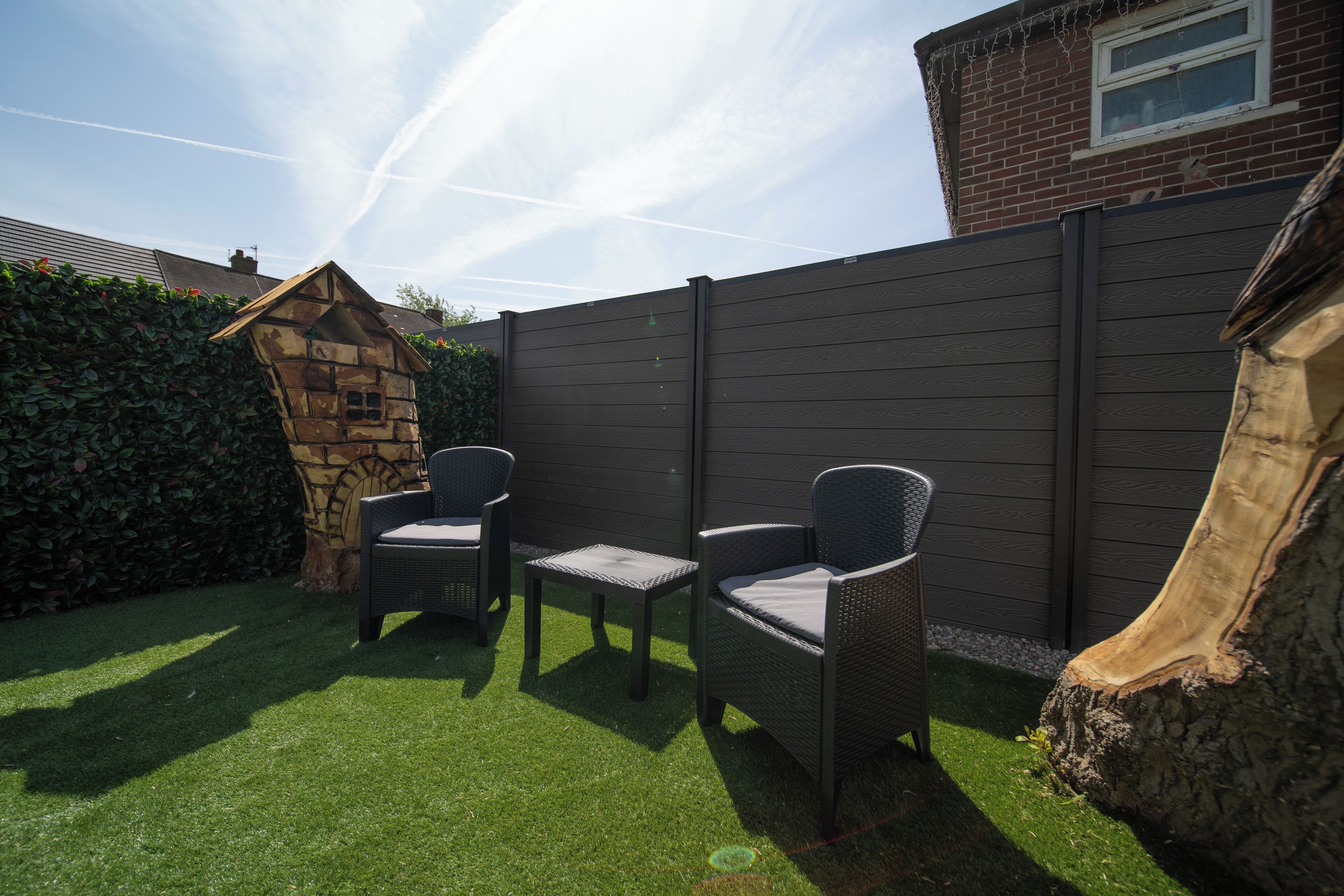 Une terrasse avec deux fauteuils en osier et une petite table, entourée d’une haie et d’une clôture en bois.