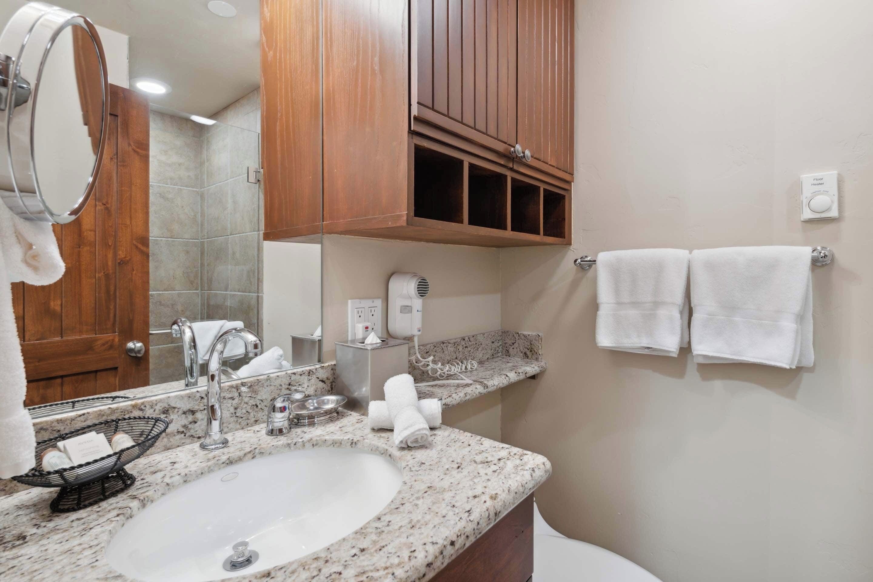 A bathroom with a granite countertop sink, a mirror, a towel rack with two towels, a soap dispenser, and a handheld showerhead.