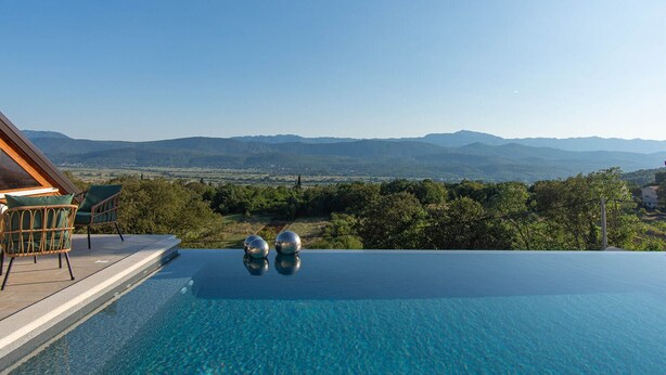 A infinity pool with a clear view of distant mountains, a wooden deck, and two wicker chairs. - Luxury Villa with big private swimming pool surrounded by forest and wineyards (Imotski)