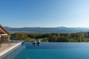A infinity pool with a clear view of distant mountains, a wooden deck, and two wicker chairs. - Luxury Villa with big private swimming pool surrounded by forest and wineyards (Imotski)