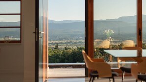 A room with a large sliding glass door leading to a scenic view of mountains and a valley. Inside, there is a table with chairs and a vase with flowers. - Luxury Villa with big private swimming pool surrounded by forest and wineyards (Imotski)