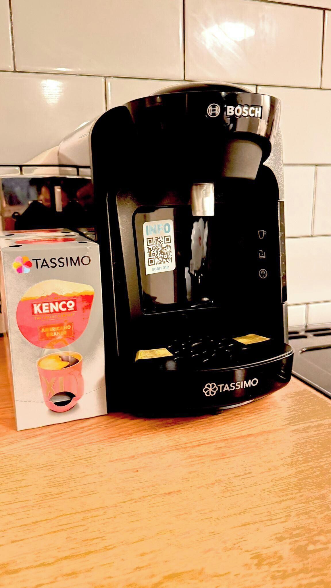 A black Tassimo coffee machine with a QR code and the Tassimo logo, placed next to its packaging.