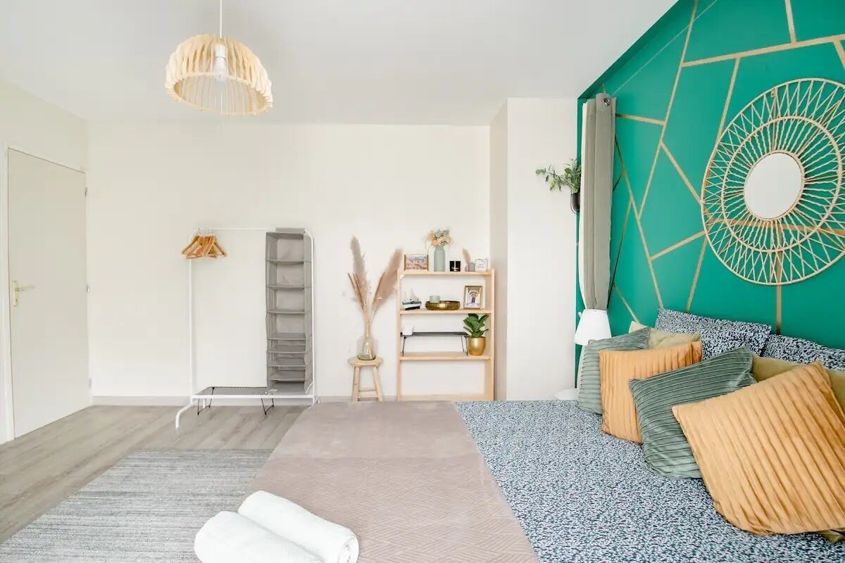 A modern bedroom with a bed, a standing lamp, a shelf with decorative items, and a geometric wall design.