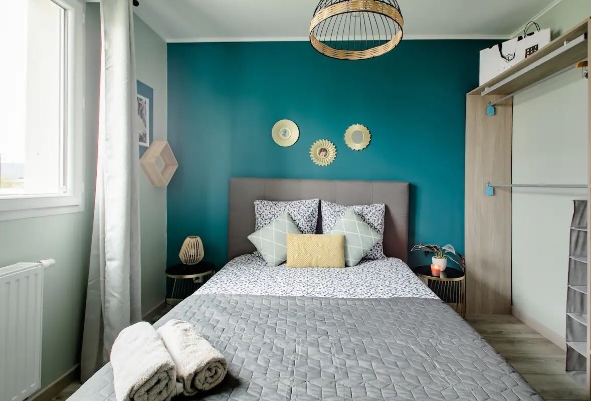 A bedroom with a bed, a headboard, a window, a wall with decorative plates, a small table with a lamp, and a wardrobe.