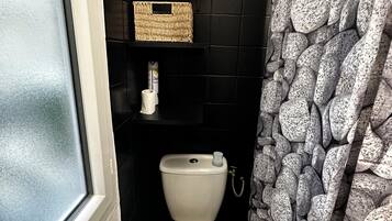A small, compact bathroom with a white toilet, a black tiled wall, a wicker basket on a shelf, and a stone-patterned shower curtain.